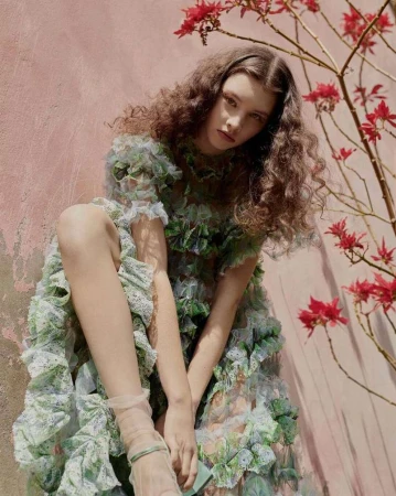 Woman with long curly brown hair wearing a ruffled tulle dress posed outdoors near a pink wall and red flowers.