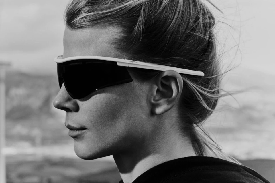 Black and white portrait of a woman in profile wearing sport sunglasses against a blurred, overcast landscape background.