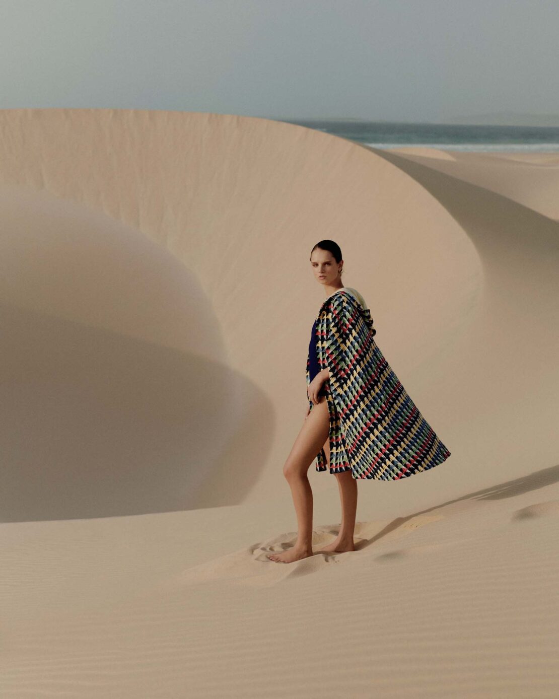 Model standing on a sandy desert dune wearing a patterned cape and dark swimsuit with the ocean visible in the background.