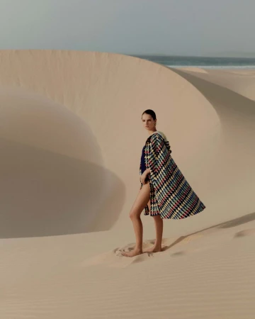 Model standing on a sandy desert dune wearing a patterned cape and dark swimsuit with the ocean visible in the background.