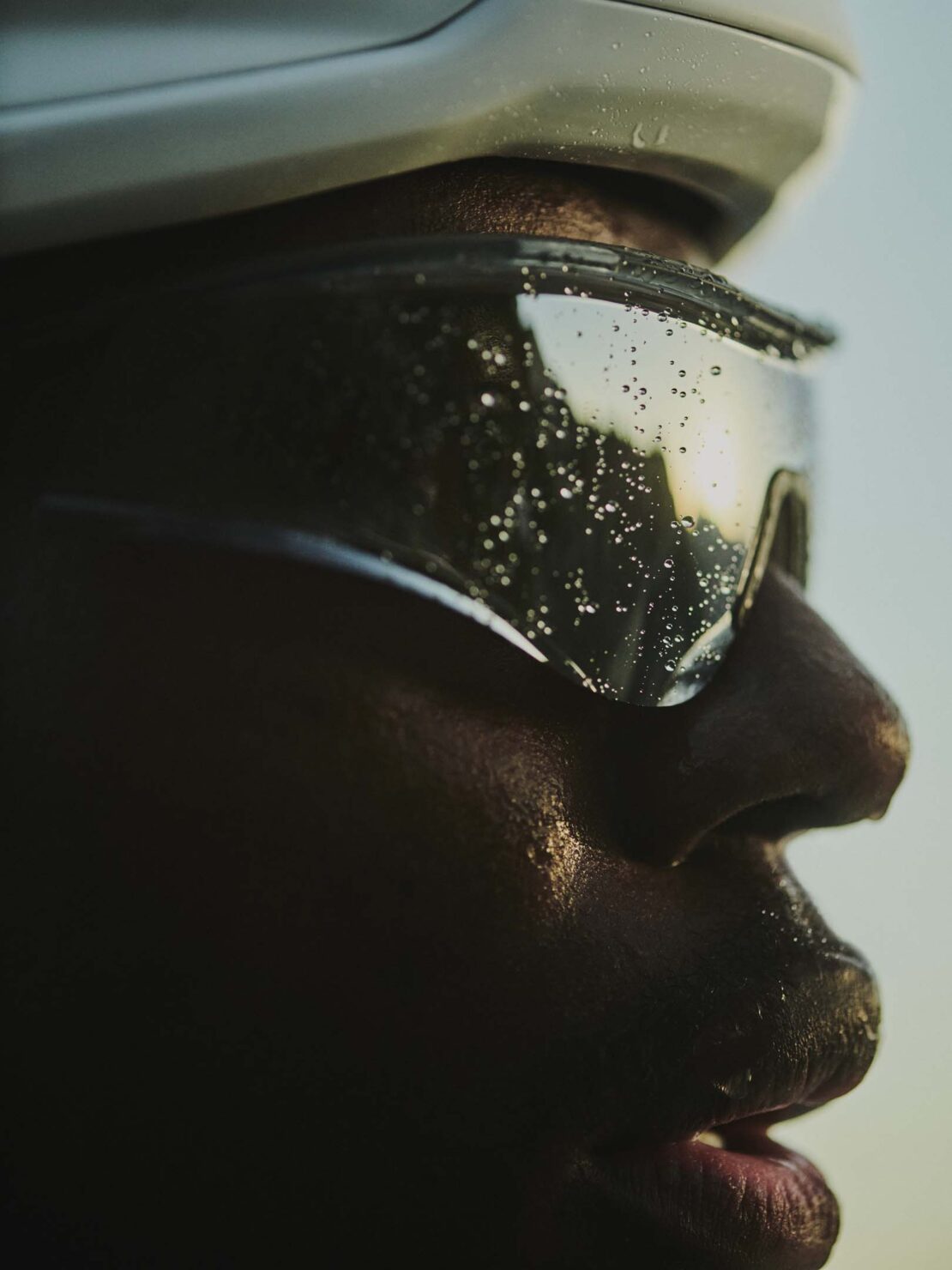 Close-up profile of a Black adult male athlete wearing a helmet and water-covered sunglasses, against a soft, light blue background.