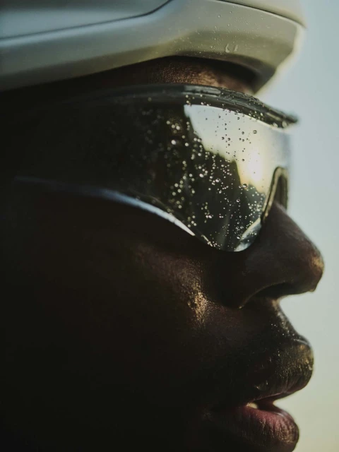 Close-up profile of a Black adult male athlete wearing a helmet and water-covered sunglasses, against a soft, light blue background.