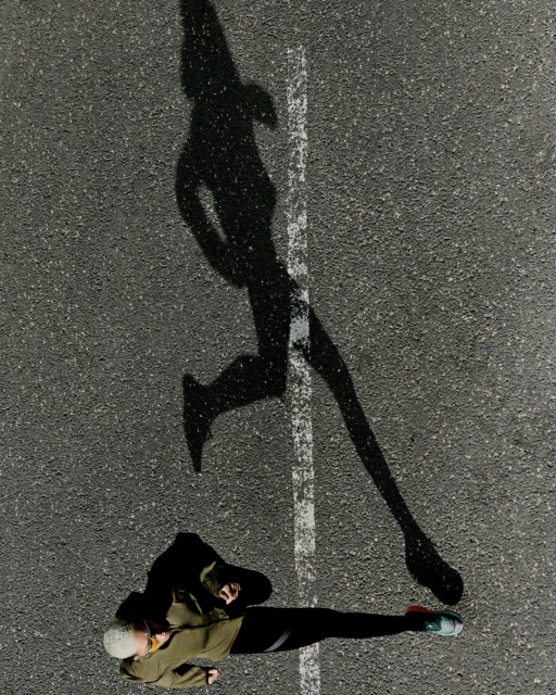 Overhead shot of a runner on an asphalt surface, casting a long shadow near a white line, emphasizing the athlete's form and motion.