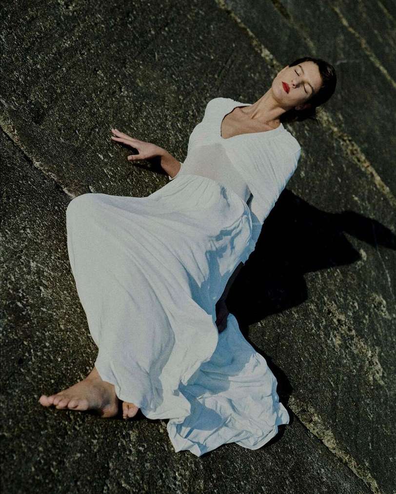 A woman in a white dress lies on a concrete surface with eyes closed, face tilted towards the sun in an outdoor setting.