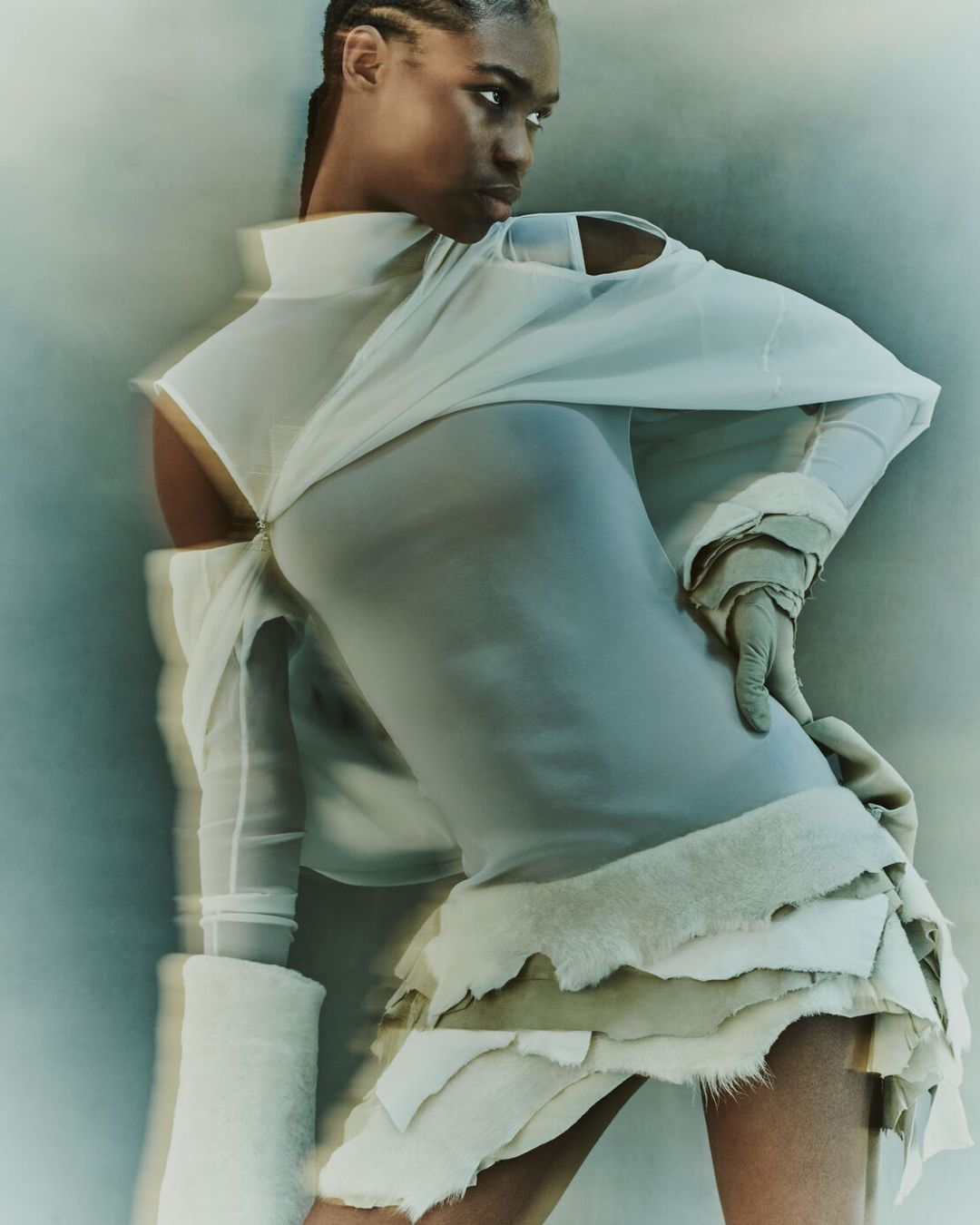 Woman in a futuristic dress with cut-out shoulders poses in a studio against a blurry background for a fashion or editorial layout.