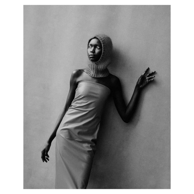 Monochrome fashion portrait of an African female model in a strapless dress and knit balaclava leaning against a textured wall in studio.