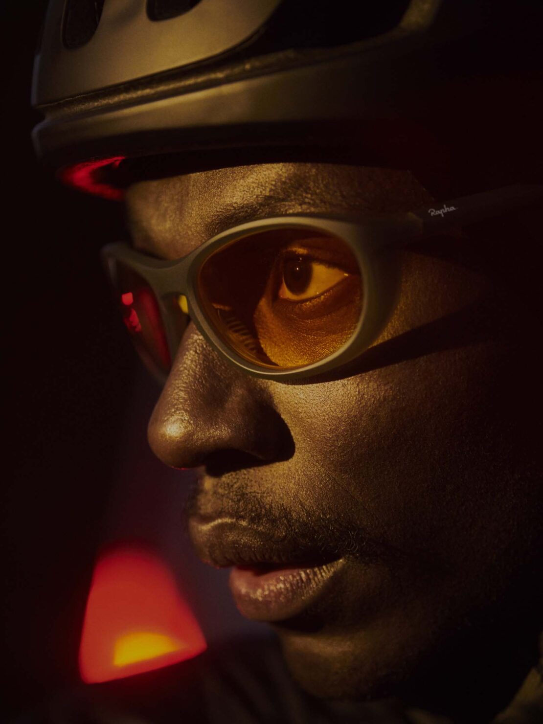 Close-up of a dark-skinned adult male wearing a cycling helmet and sunglasses, illuminated by warm, directional lighting in a dark setting.