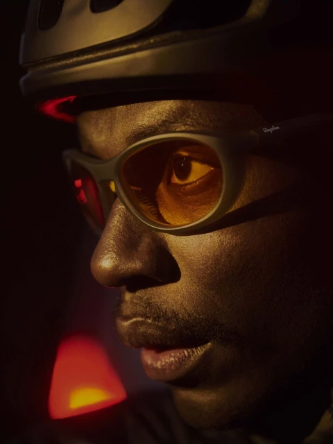 Close-up of a dark-skinned adult male wearing a cycling helmet and sunglasses, illuminated by warm, directional lighting in a dark setting.