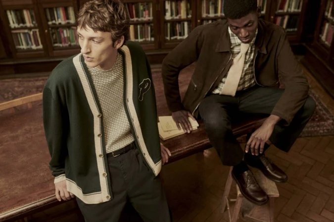 Two stylish men in vintage-inspired outfits in a library setting, one in a varsity cardigan, the other in a brown jacket and tie.
