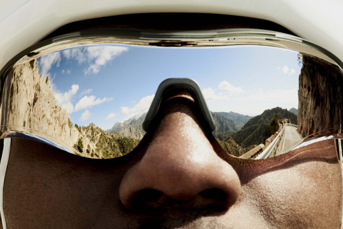 Close-up POV perspective of adult person wearing mirrored visor reflecting mountain road landscape and blue sky with white clouds.