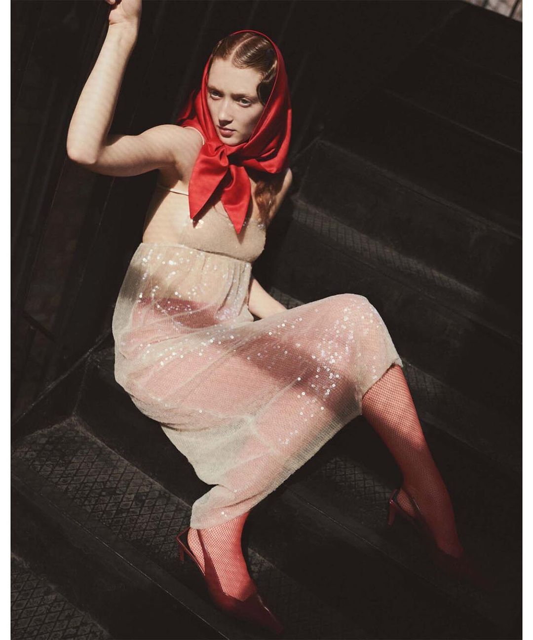 Woman in a sheer beige dress with glitter and a red headscarf poses on a black staircase in soft, diffused sunlight indoors.