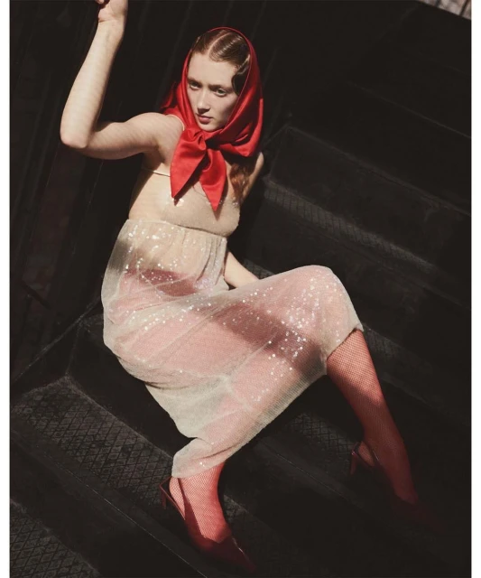 Woman in a sheer beige dress with glitter and a red headscarf poses on a black staircase in soft, diffused sunlight indoors.
