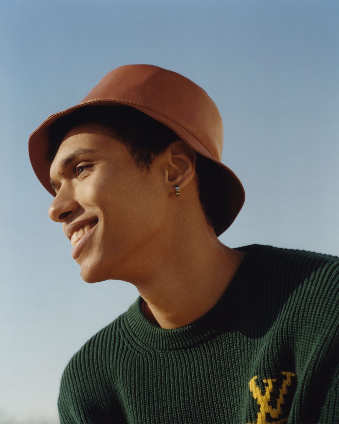 Portrait of an adult male with a smile wearing a brown bucket hat and a green sweater with a yellow pattern against a blue sky.