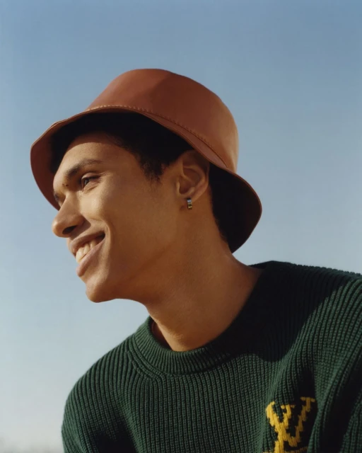 Portrait of an adult male with a smile wearing a brown bucket hat and a green sweater with a yellow pattern against a blue sky.