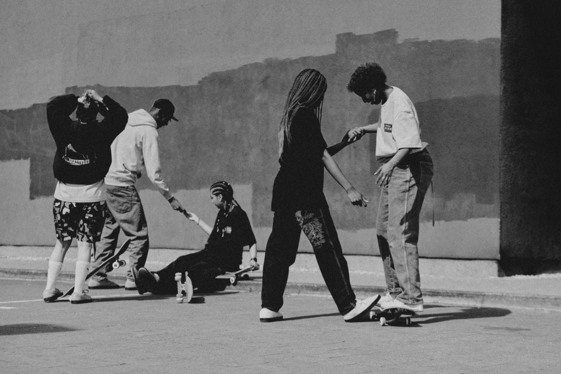 Black and white photograph of a group of young adult skateboarders, some helping others, posing together on the street near a stucco wall.