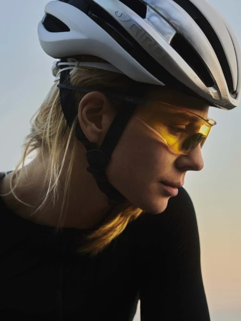 Close-up of a woman wearing a white bicycle helmet and yellow-tinted sports glasses, with a blurred gradient sky in the background.
