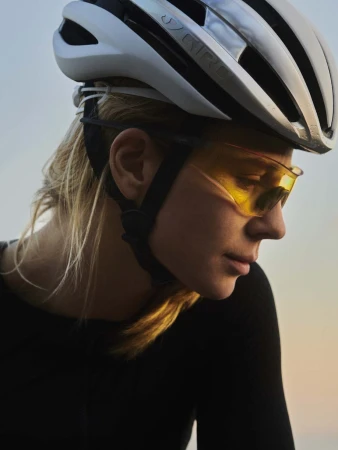 Close-up of a woman wearing a white bicycle helmet and yellow-tinted sports glasses, with a blurred gradient sky in the background.