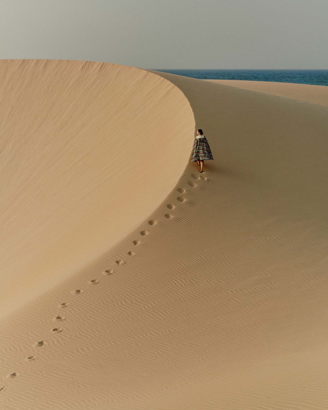 Woman in patterned shawl walking on sand dune with ocean view. Footprints visible in sand. Tan, gray, and blue tones. Desert landscape.