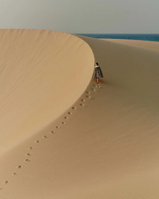 Woman in patterned shawl walking on sand dune with ocean view. Footprints visible in sand. Tan, gray, and blue tones. Desert landscape.
