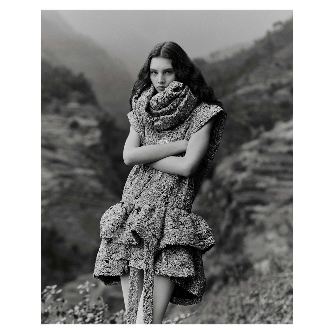 Monochrome fashion portrait of a woman in a textured dress and scarf, with a mountain landscape backdrop, and a neutral expression.