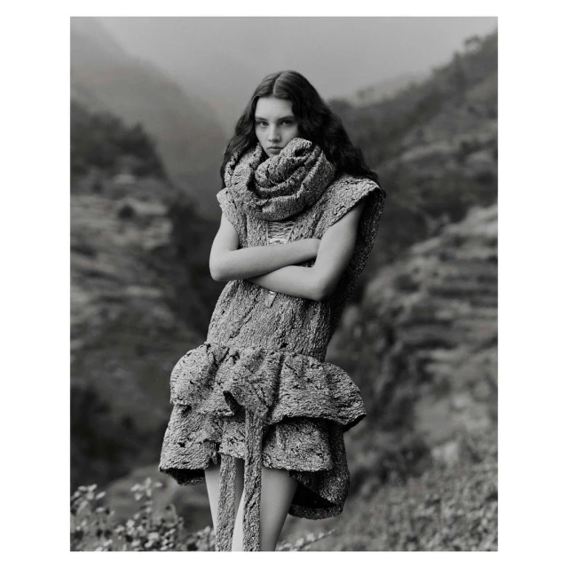 Monochrome fashion portrait of a woman in a textured dress and scarf, with a mountain landscape backdrop, and a neutral expression.