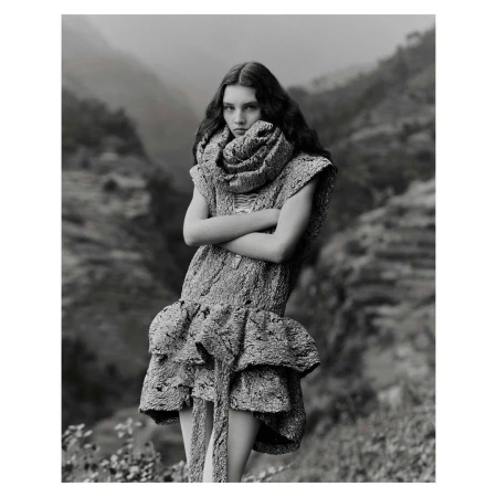 Monochrome fashion portrait of a woman in a textured dress and scarf, with a mountain landscape backdrop, and a neutral expression.