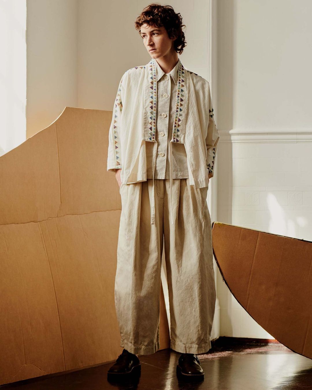 Full-length portrait of a young adult woman in a patterned beige pantsuit, standing in a room with large cardboard cutouts.