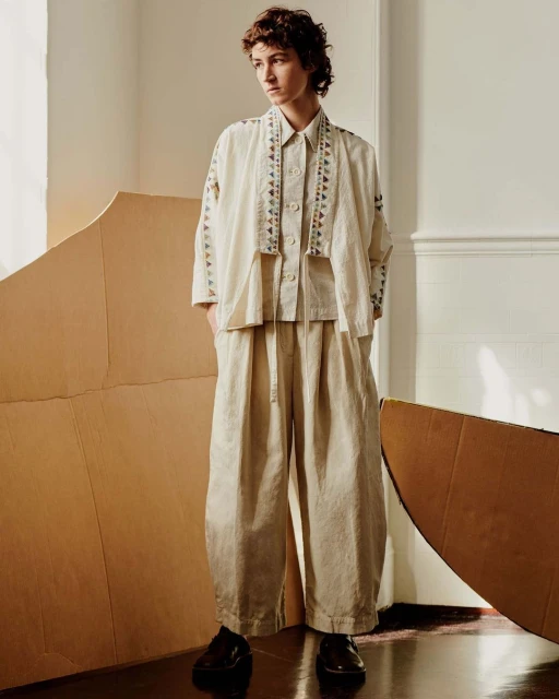 Full-length portrait of a young adult woman in a patterned beige pantsuit, standing in a room with large cardboard cutouts.
