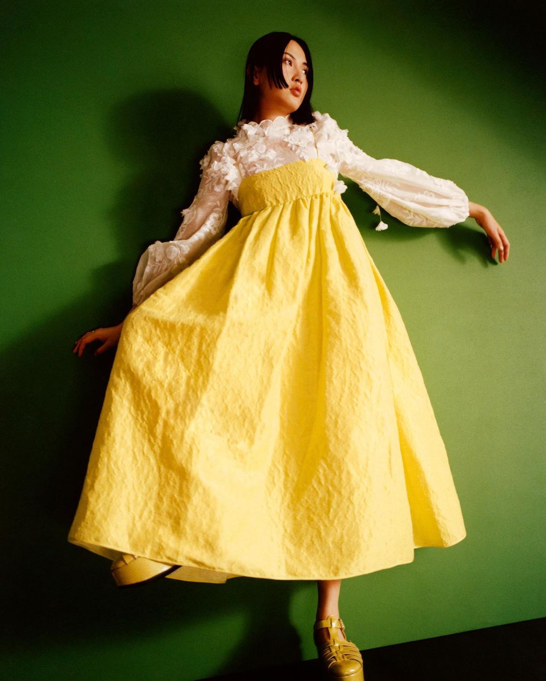 Portrait of a young Asian woman in a yellow dress with white floral top against a green background, looking off camera with a slight smile.