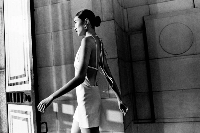 Monochrome fashion portrait of an Asian woman walking in a short dress, featuring architectural details and a minimalist aesthetic.