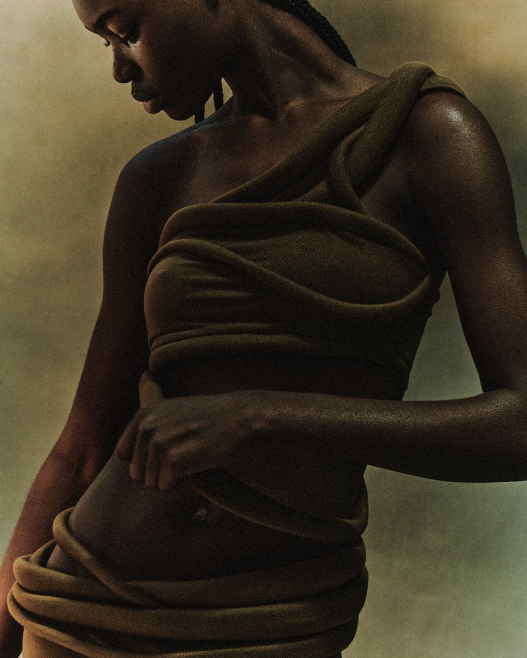 A woman with dark skin wears a draped, olive-green fabric dress in a studio portrait. The dress is wrapped around her body.