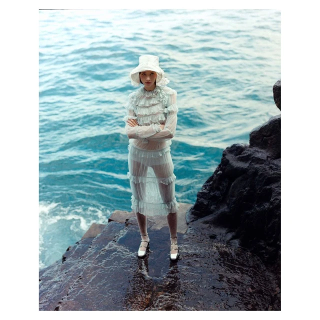 A lone woman on a rocky shore stands facing the camera in a sheer ruffled dress and bucket hat, with the ocean as her backdrop.
