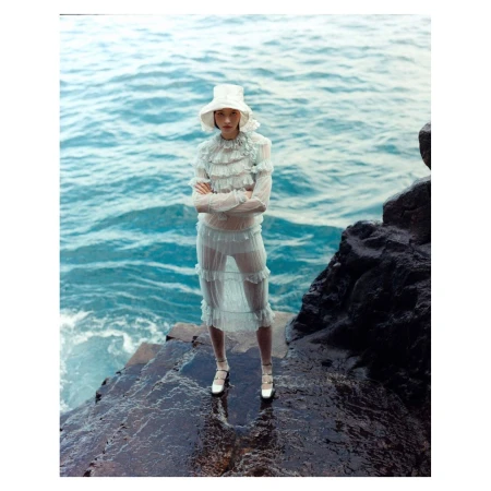 A lone woman on a rocky shore stands facing the camera in a sheer ruffled dress and bucket hat, with the ocean as her backdrop.