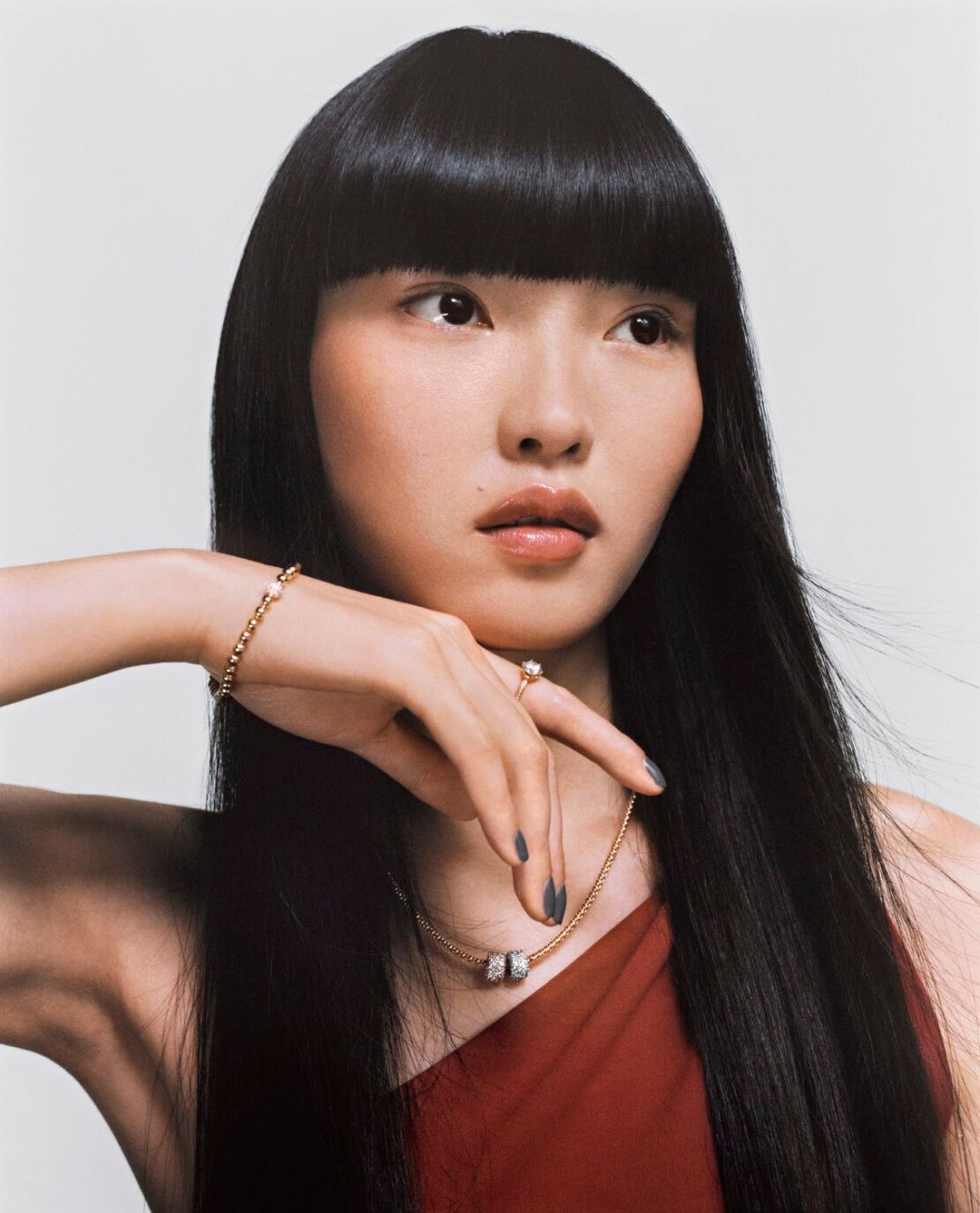 Portrait of a woman with long black hair wearing gold jewellery and a red one-shoulder top against a neutral background
