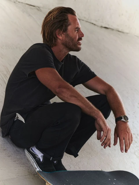 Adult male with long hair in black t-shirt crouching in concrete skatepark with skateboard and watch in medium shot, side view.