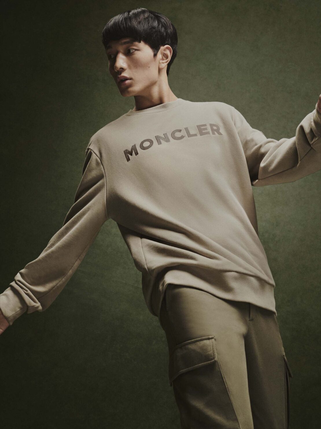 Asian male model wearing a Moncler sweatshirt and pants in a studio setting with a green background, fashion editorial style.