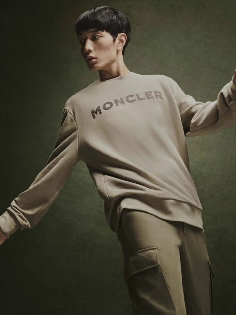 Asian male model wearing a Moncler sweatshirt and pants in a studio setting with a green background, fashion editorial style.