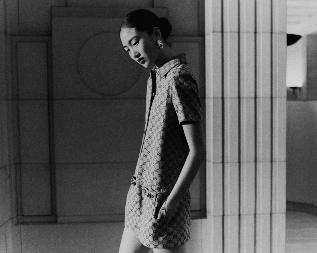 Monochrome shot of a stylish Asian woman in a patterned dress with hand in pocket, captured indoors with architectural details.