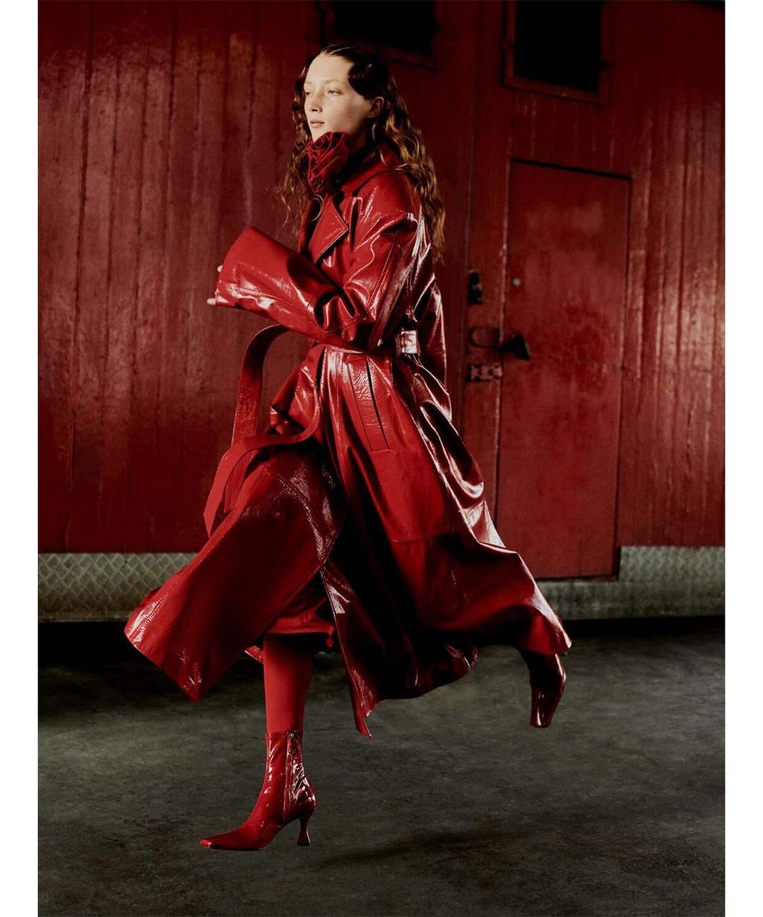 A woman in a shiny red coat and matching boots poses in front of a red wall, creating a striking, monochromatic fashion statement.