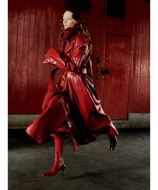 A woman in a shiny red coat and matching boots poses in front of a red wall, creating a striking, monochromatic fashion statement.