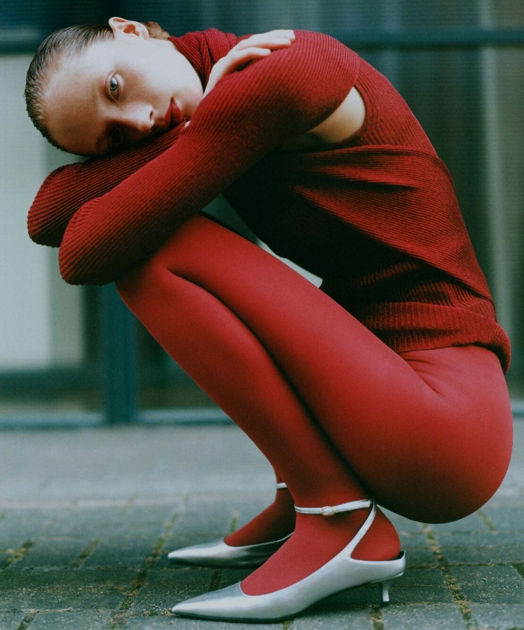 Woman in a red ribbed turtleneck and red tights crouching outdoors on stone paving wearing silver kitten heels
