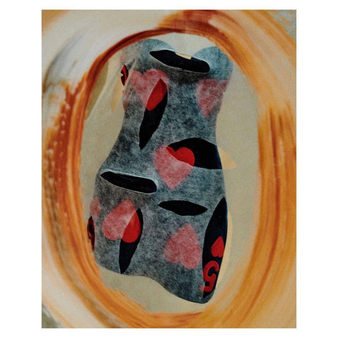 Abstract art featuring a dress form shape with red heart and black oval patterns on a textured grey background encircled by orange paint.