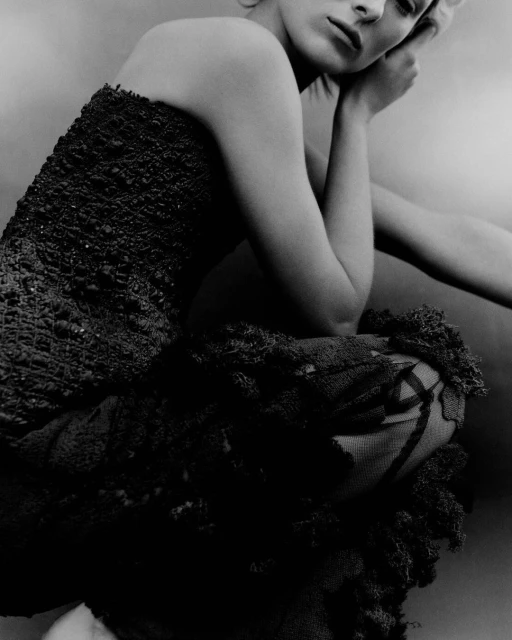 Monochrome portrait of an adult female fashion model crouching with hands on face and wearing a textured strapless dress in a studio.