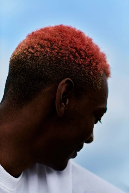Close-up of an adult male with short, red-tinted hair. His head is turned in profile against a light blue sky, with a white top visible.