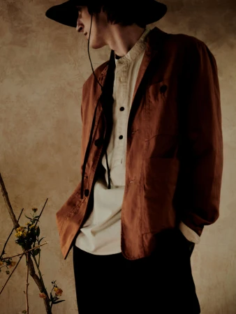Portrait of a man in a rust-colored jacket, white button-down shirt, and black hat against a textured stone wall
