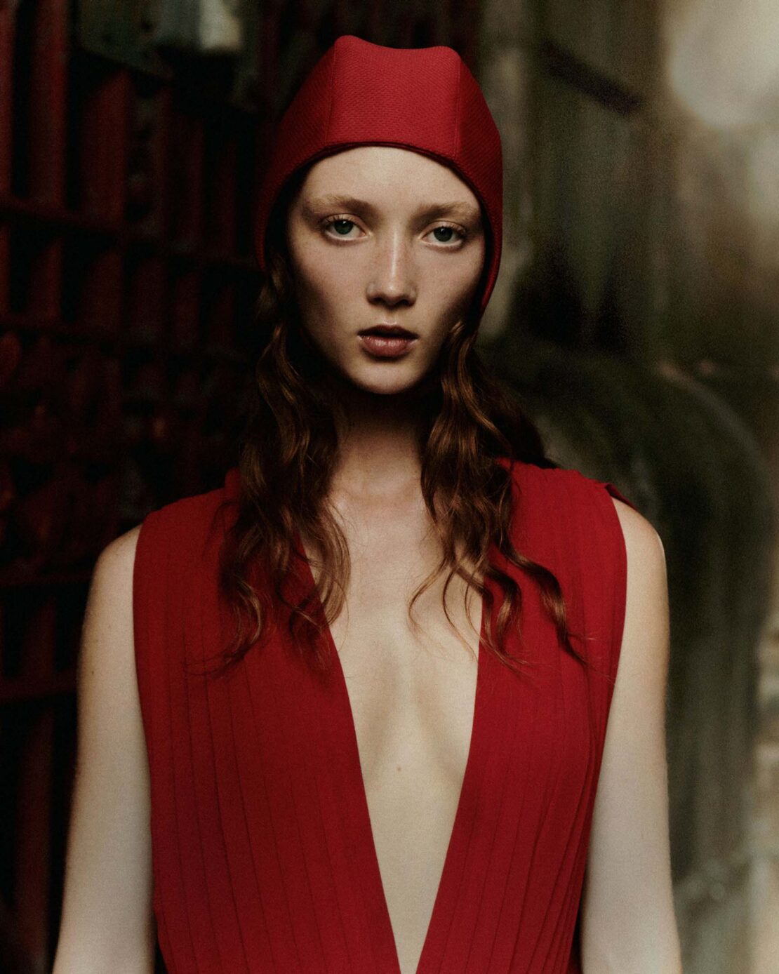 Portrait of a fair-skinned adult woman with auburn hair wearing a red cap and red v-neck dress, posing in front of a dark stone wall.