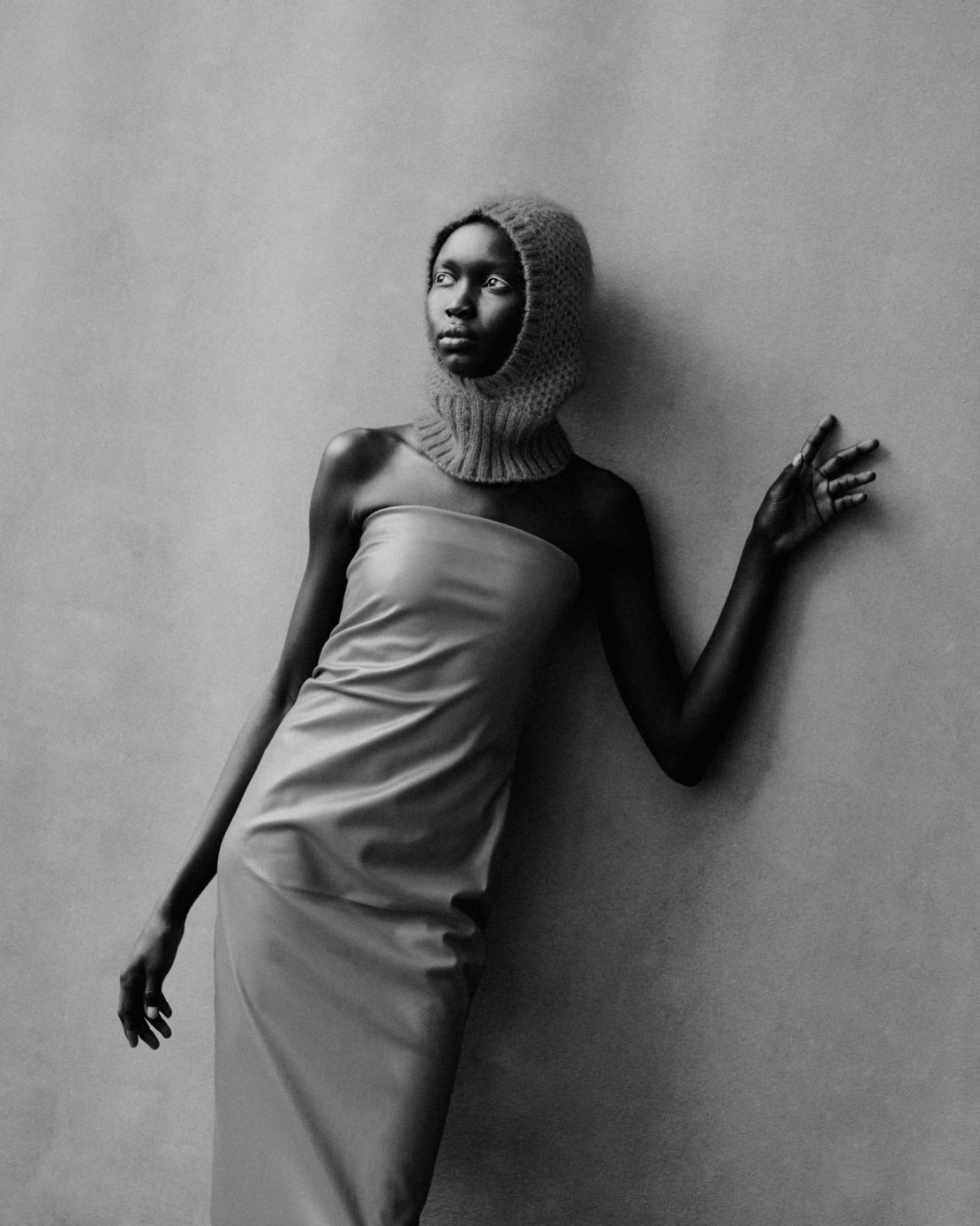Elegant black and white studio portrait of a woman in a textured hood and a draped dress, gazing off-camera in a fashion-forward pose.