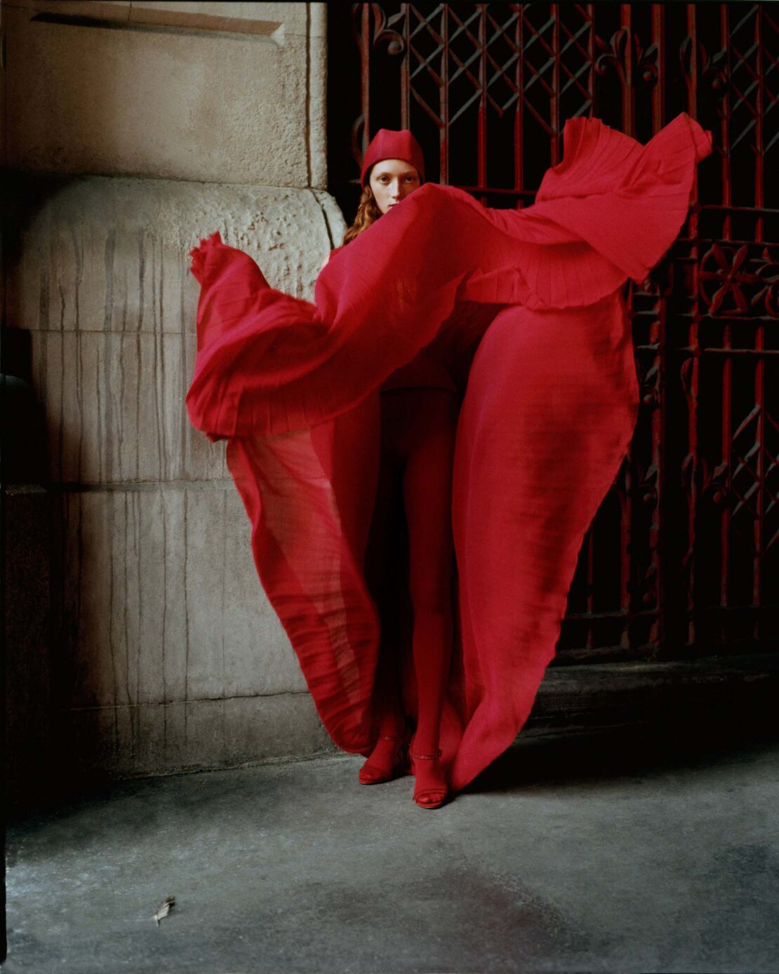 Fashion shot of a woman in a red outfit posing in a flowing red dress, matching red head covering and red heels, with a red metal gate.