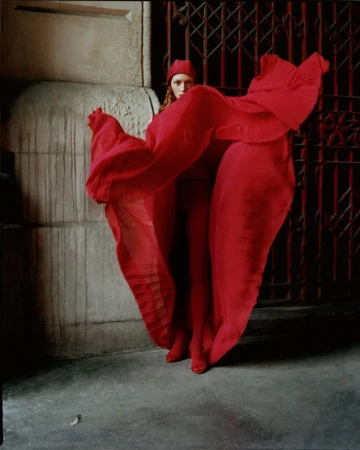 Fashion shot of a woman in a red outfit posing in a flowing red dress, matching red head covering and red heels, with a red metal gate.