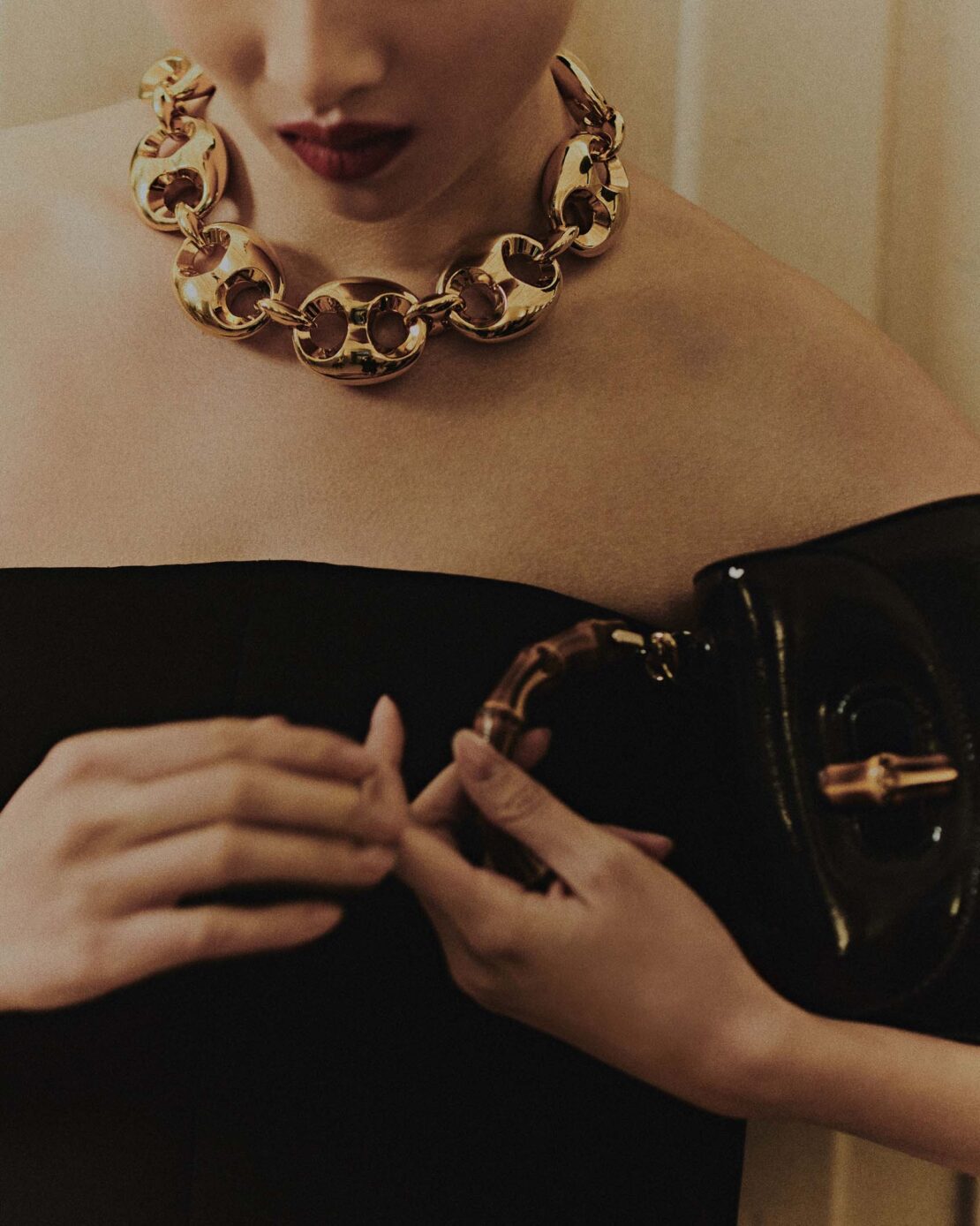 Elegant woman showcasing a gold chain necklace and a black patent leather handbag, highlighted by indoor studio lighting.